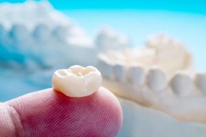 Dental crown resting on a fingertip in front of model jaws and teeth